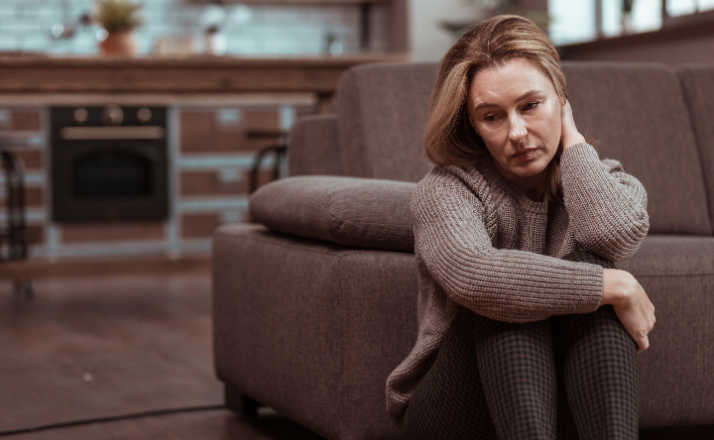 Woman wearing brown squared trousers feeling depressed sitting on the floor next to her couch. This is depression