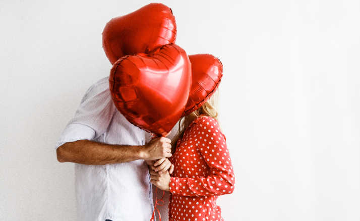 Couple. Love. Valentine's day. Emotions. Man is giving heart-shaped balloons to his woman, both smiling; on a white background