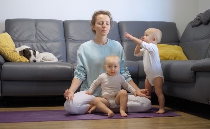 Mom in yoga pants closing her eyes as twin babies crawl around her