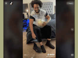Screenshot of a man sitting on a bed with a watermelon plastic wrapped to his belly.