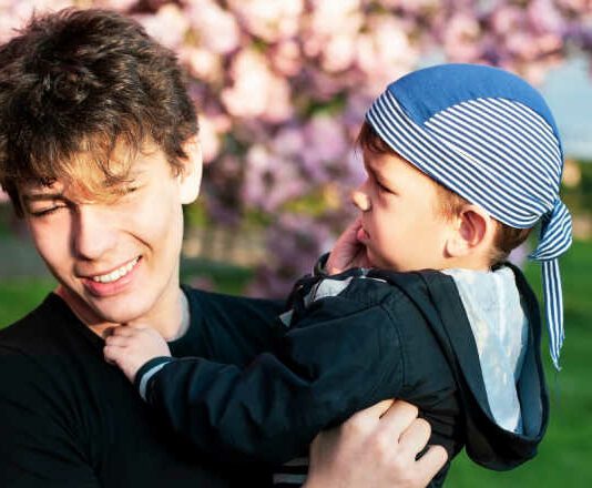 Two brothers - a young boy and a teen together fooling around during a photo shoot against the background of sakura blossoms with pink flowers. The brothers are friends and look after each other