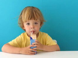 Cute focused boy in yellow t-shirt drinking from carton box through straw sitting on blue background