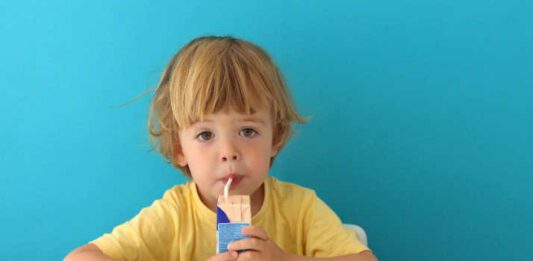 Cute focused boy in yellow t-shirt drinking from carton box through straw sitting on blue background