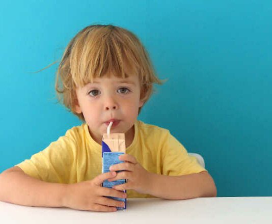 Cute focused boy in yellow t-shirt drinking from carton box through straw sitting on blue background