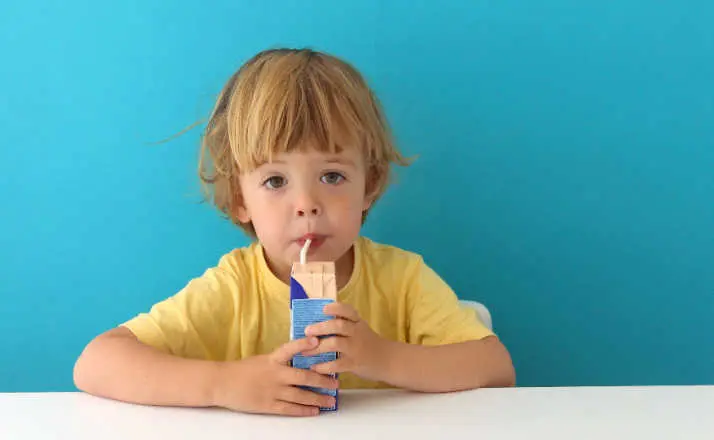 Cute focused boy in yellow t-shirt drinking from carton box through straw sitting on blue background