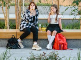 Two schoolgirls, a child and a teenager, have their lunch on a bench in the schoolyard.