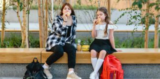 Two schoolgirls, a child and a teenager, have their lunch on a bench in the schoolyard.