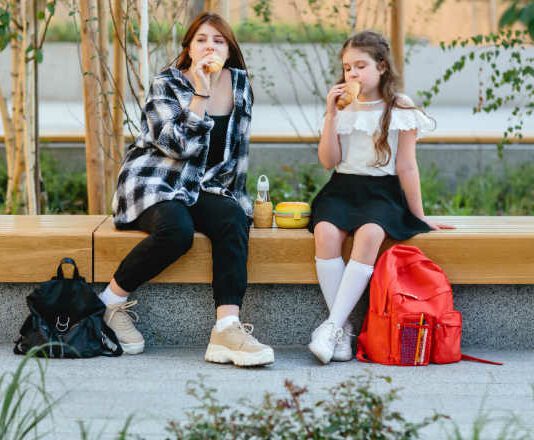 Two schoolgirls, a child and a teenager, have their lunch on a bench in the schoolyard.