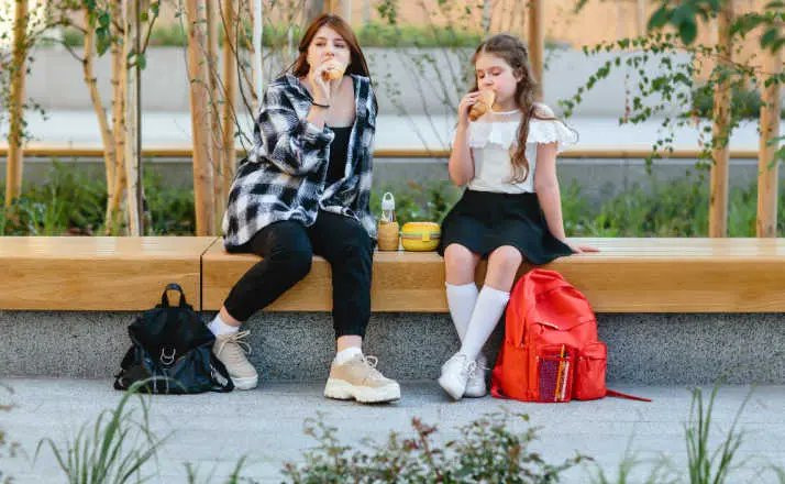 Two schoolgirls, a child and a teenager, have their lunch on a bench in the schoolyard.