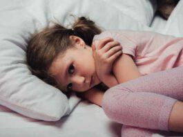 frightened child looking at camera while lying in bed and hugging herself with arms