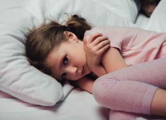 frightened child looking at camera while lying in bed and hugging herself with arms