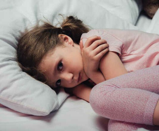frightened child looking at camera while lying in bed and hugging herself with arms