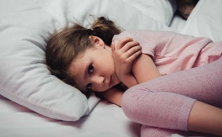 frightened child looking at camera while lying in bed and hugging herself with arms