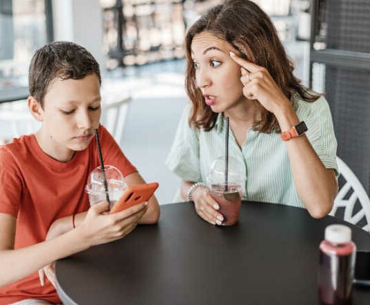 Emotional conflict between mother and son about smartphone addiction in cafe. An angry mom and a teenager ignoring her.