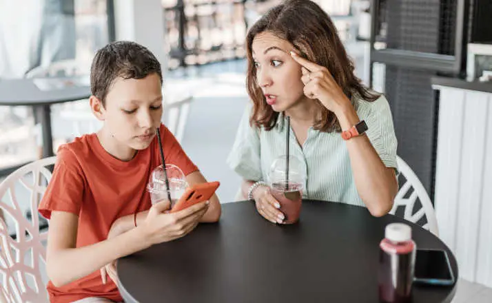 Emotional conflict between mother and son about smartphone addiction in cafe. An angry mom and a teenager ignoring her.