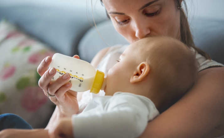 Mother, feeding her baby boy from bottle, comfortably sitting on the couch at home