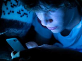 young boy using a smartphone lying in bed under a blanket at night