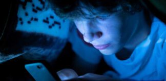 young boy using a smartphone lying in bed under a blanket at night