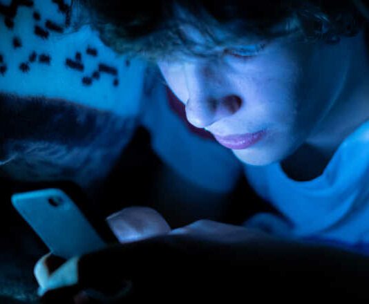 young boy using a smartphone lying in bed under a blanket at night