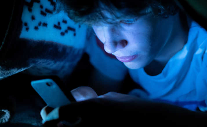 young boy using a smartphone lying in bed under a blanket at night