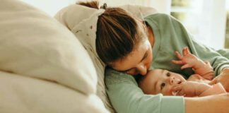 Young mother taking a nap with her arms around her baby. New mom resting while her baby plays with a pacifier. Mother and baby lying on the bed together at home.