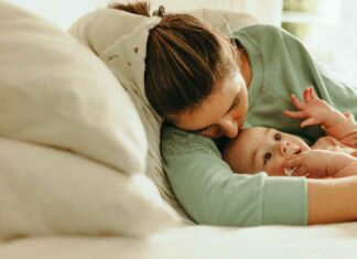 Young mother taking a nap with her arms around her baby. New mom resting while her baby plays with a pacifier. Mother and baby lying on the bed together at home.