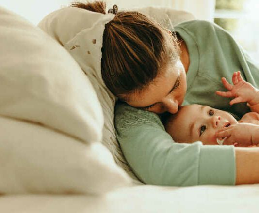 Young mother taking a nap with her arms around her baby. New mom resting while her baby plays with a pacifier. Mother and baby lying on the bed together at home.