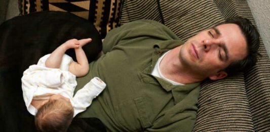 A newborn baby is curled up asleep on her sleeping father.