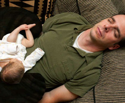 A newborn baby is curled up asleep on her sleeping father.