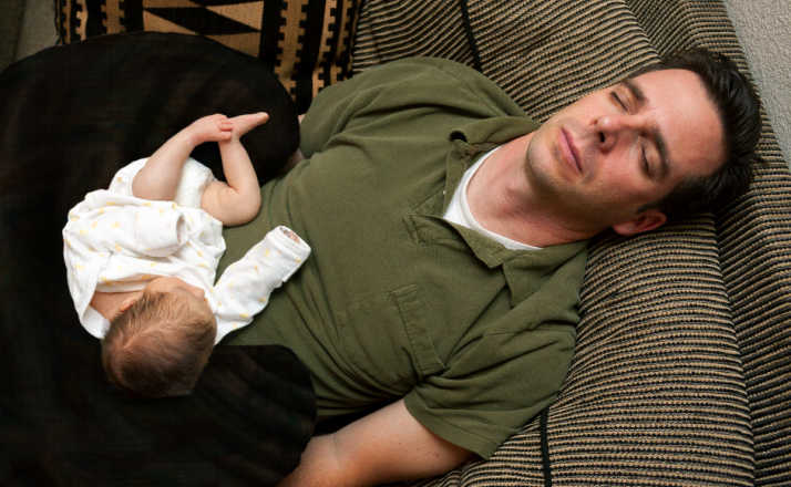 A newborn baby is curled up asleep on her sleeping father.