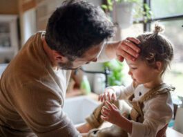 flu season Father checking forehead of sick toddler daughter indoors in kitchen at home.