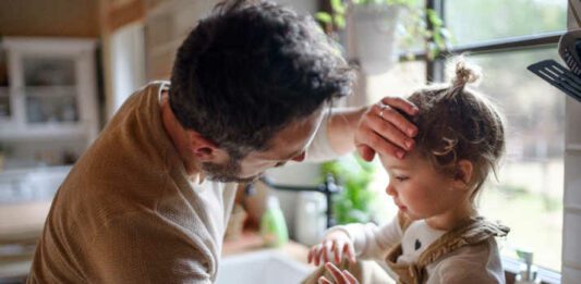 flu season Father checking forehead of sick toddler daughter indoors in kitchen at home.