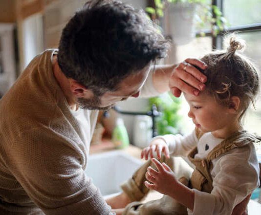 flu season Father checking forehead of sick toddler daughter indoors in kitchen at home.